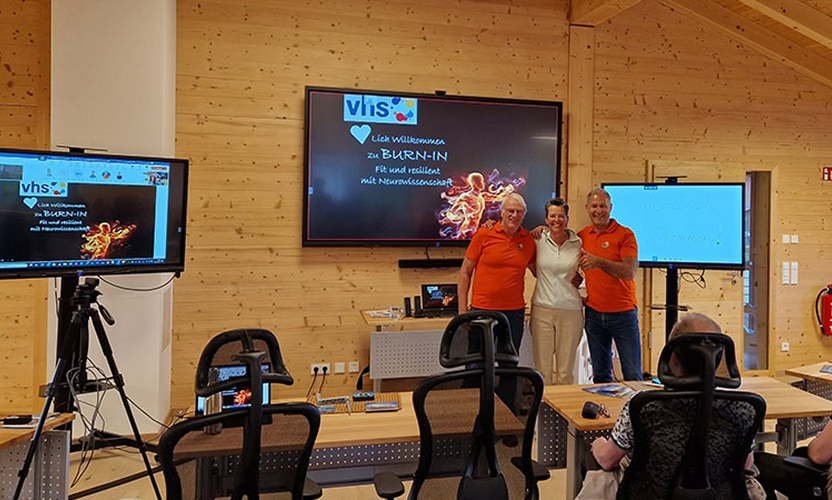 Drei Personen in orangefarbenen T-Shirts stehen vor Bildschirmen in einem Seminarraum für das BURN-IN Programm.