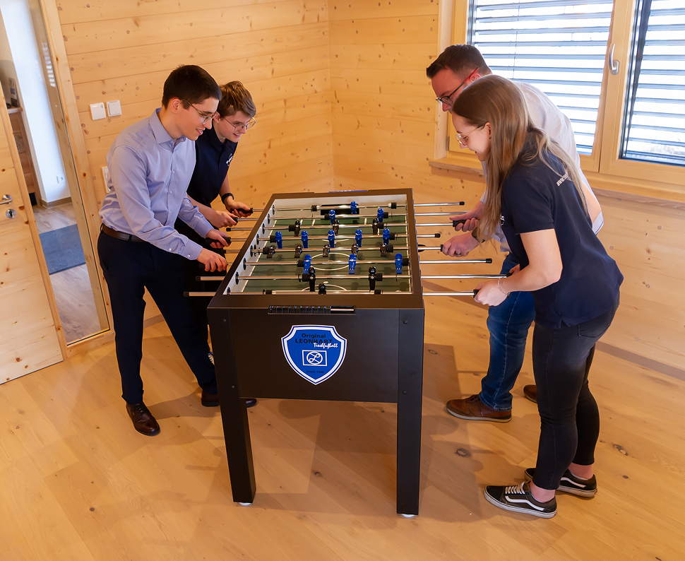 Vier Personen spielen an einem Kickertisch in einem Raum mit Holzverkleidung und großen Fenstern.