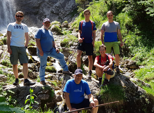Sechs Personen wandern in den Bergen nahe eines Wasserfalls in einer natürlichen Landschaft.