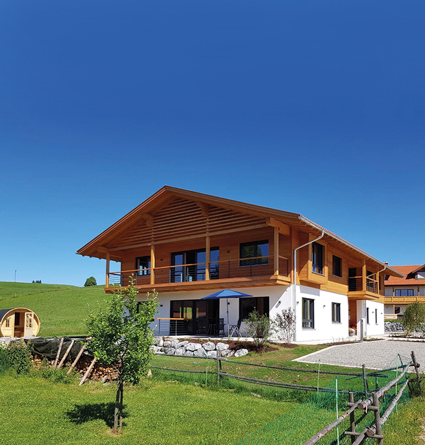 Zweistöckiges Gebäude mit Holzfassade und Balkon in einer Wiesenlandschaft. Links steht ein kleines Holzfass.