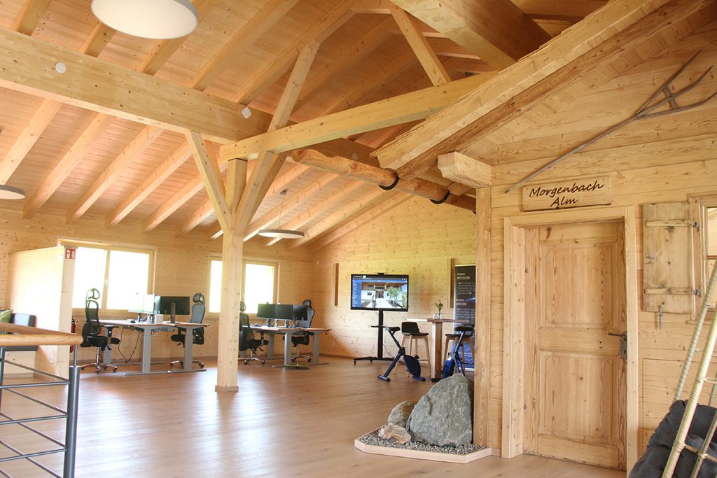 Großer Arbeitsraum aus hellem Holz mit mehreren Schreibtischen und Monitoren. Rechts steht eine kleine Holzhütte mit dem Schild „Morgenbach Alm“.