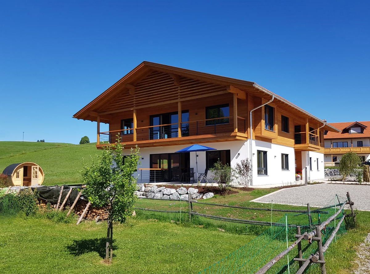 Modernes Holzhaus mit Balkon, Garten und Blick auf die umliegende Landschaft in sonniger Umgebung. Ideal f&uuml;r Erholung.