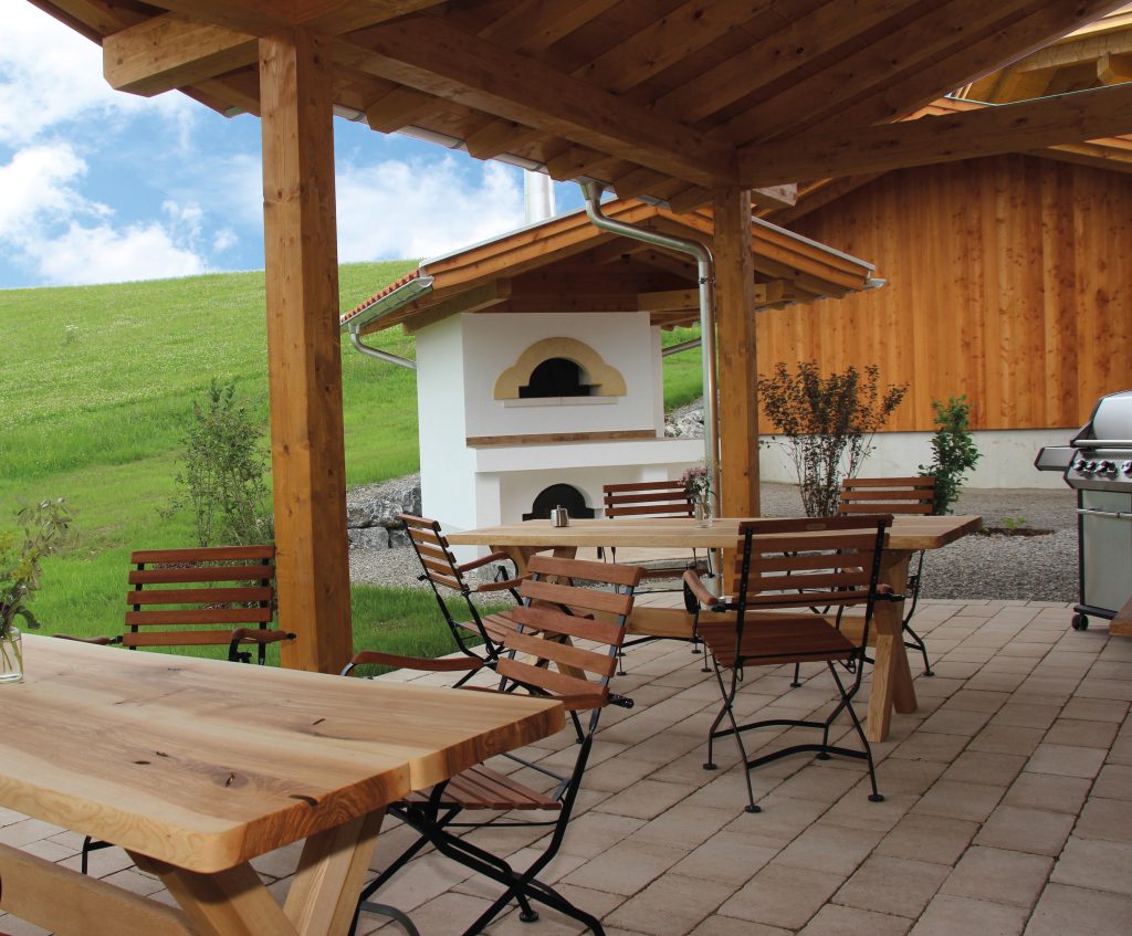 Außenbereich mit Holzdach, Tischen und Stühlen, einem Grill und einem Steinofen, umgeben von Wiesen und Holzfassaden.
