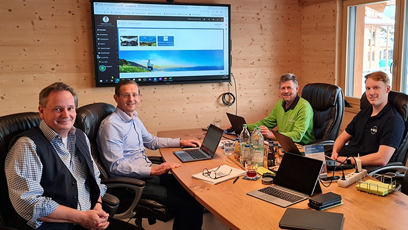 Vier Personen sitzen an einem Tisch mit Laptops. Im Hintergrund läuft eine Präsentation auf einem Bildschirm. Der Raum besteht aus hellem Holz.